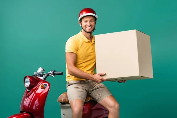 Hombre de entrega feliz en casco celebración de caja de cartón grande cerca de scooter en azul - foto de stock