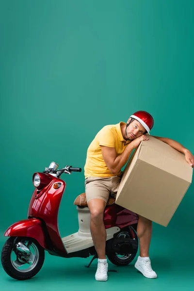 Tired delivery man in helmet holding big carton box near scooter on blue — Stock Photo