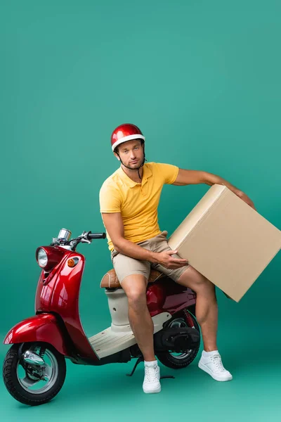 Homem de entrega exausto no capacete segurando grande caixa de papelão perto de scooter em azul — Fotografia de Stock