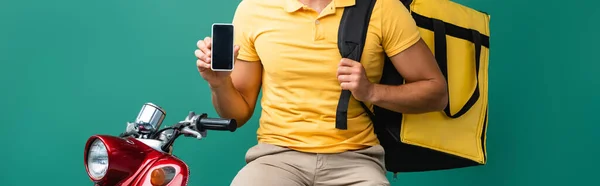 Vista parcial del repartidor con la mochila que sostiene el teléfono inteligente con la pantalla en blanco cerca del scooter en azul - foto de stock
