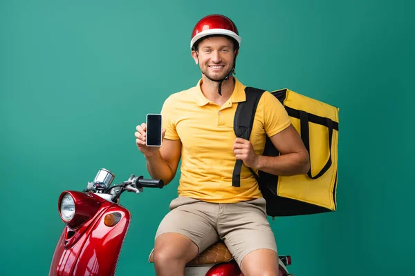 Щасливий чоловік доставки з рюкзаком тримає смартфон з порожнім екраном біля скутера на синьому — стокове фото