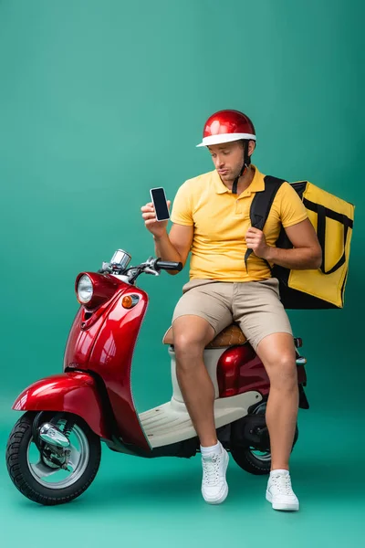 Sad delivery man with backpack holding smartphone with blank screen near scooter on blue — Stock Photo