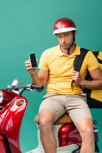 Upset delivery man with backpack holding smartphone with blank screen near scooter on blue — Stock Photo