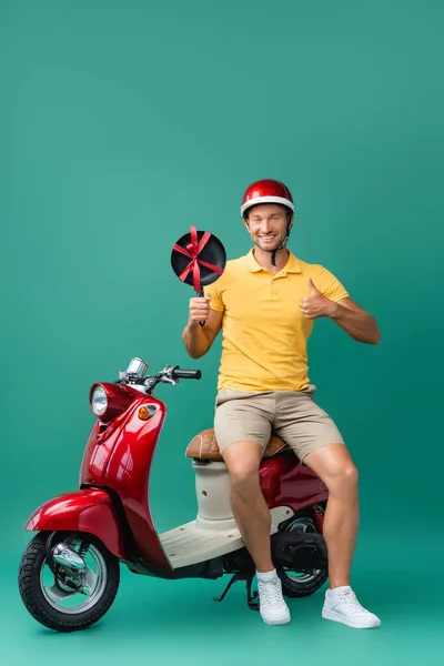 Homem de entrega feliz no capacete segurando nova frigideira e mostrando polegar perto de scooter em azul — Fotografia de Stock