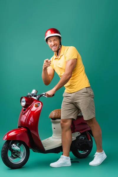 Happy delivery man pointing with finger and winking eye near scooter on blue — Stock Photo
