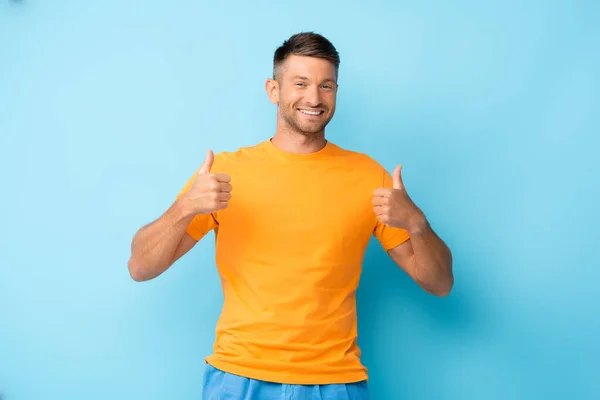 Hombre feliz en camiseta amarilla mostrando pulgares hacia arriba en azul - foto de stock