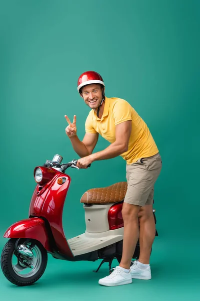 Happy delivery man in helmet standing near red scooter and showing peace sign on blue — Stock Photo