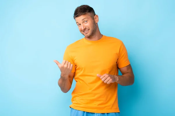 Cheerful man in yellow t-shirt pointing with thumbs on blue — Stock Photo