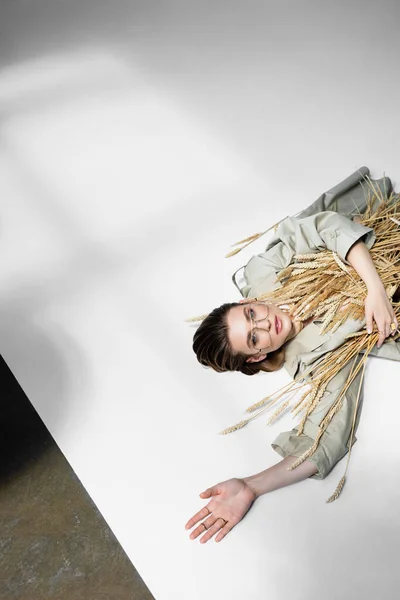 Top view of young trendy woman in glasses lying near bunch of wheat on white — Stock Photo