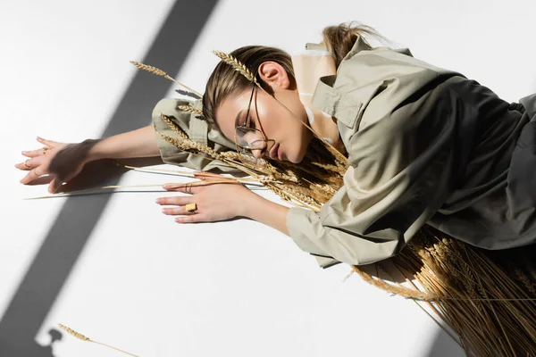 Jovem mulher na moda em óculos deitado com um monte de espiguetas de trigo no branco — Fotografia de Stock