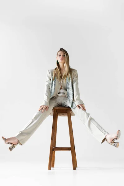 Trendy woman in suit sitting on wooden stool and looking at camera on white — Stock Photo