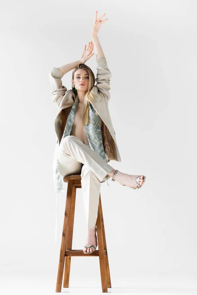 Mujer de moda en traje sentado en taburete de madera con las manos por encima de la cabeza en blanco - foto de stock