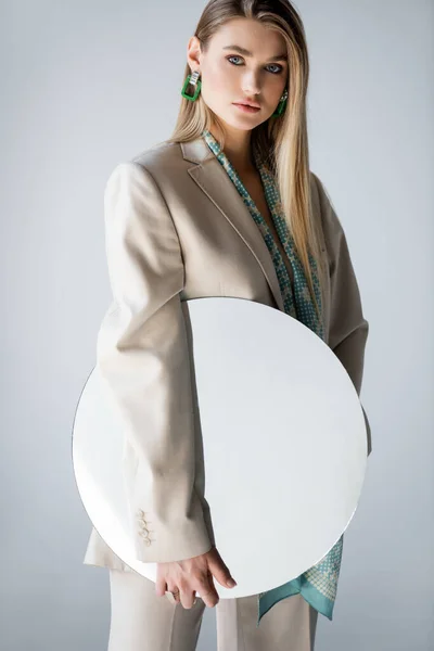 Mujer con estilo en traje sosteniendo espejo redondo mientras mira a la cámara en gris - foto de stock