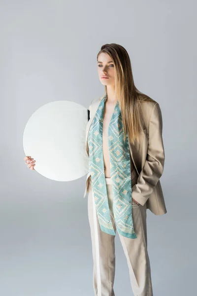 Young woman in suit holding round mirror while standing with hand in pocket on grey — Stock Photo