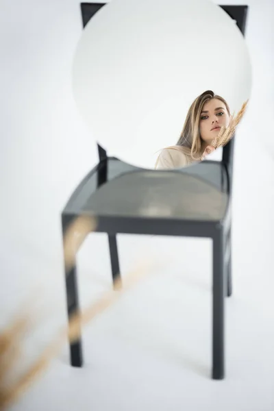 Reflexão da jovem mulher olhando para a câmera perto de trigo com cadeira borrada no fundo branco — Fotografia de Stock