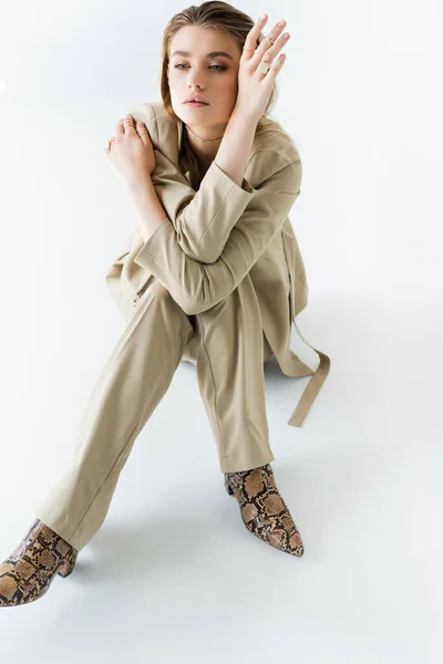 Modelo elegante em terno bege e botas com impressão animal sentado em branco — Fotografia de Stock