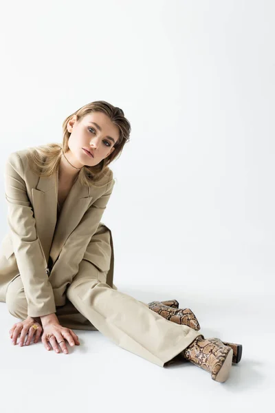 Modelo de moda en traje beige y botas con estampado animal sentado en blanco - foto de stock