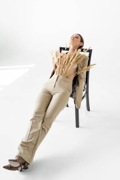 Стильная модель в бежевом костюме с пшеницей позирует на стуле на белом фоне — стоковое фото