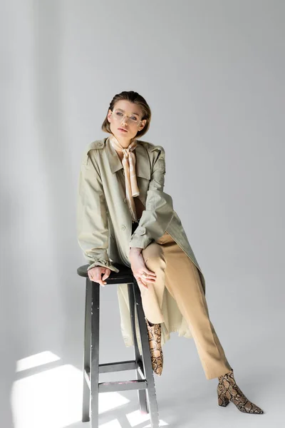 Full length of young woman in trench coat and scarf sitting on stool while posing on grey — Stock Photo