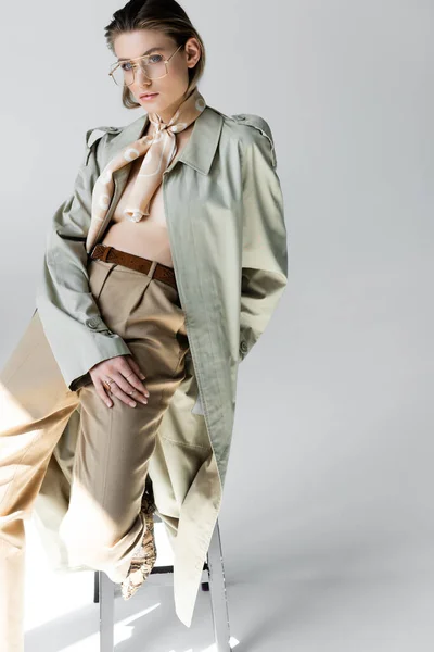 Jovem mulher na moda em casaco de trincheira e cachecol sentado nas fezes enquanto posando em cinza — Fotografia de Stock