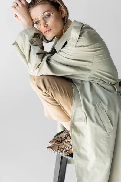 Élégant jeune modèle en trench coat, lunettes et écharpe assis sur chaise isolé sur blanc — Photo de stock