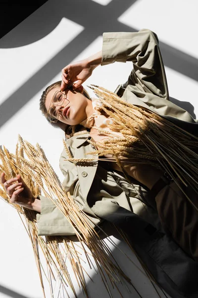 Vista aerea di giovane donna in occhiali, trench e sciarpa sdraiata vicino al grano sul bianco — Stock Photo