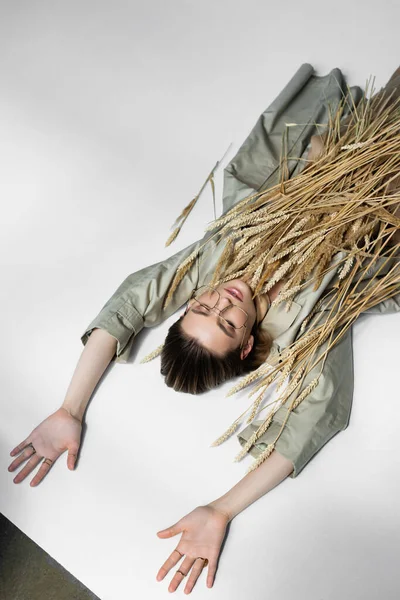 Vista ad alto angolo della donna in occhiali, trench e sciarpa sdraiato vicino a spikelets grano su bianco — Foto stock