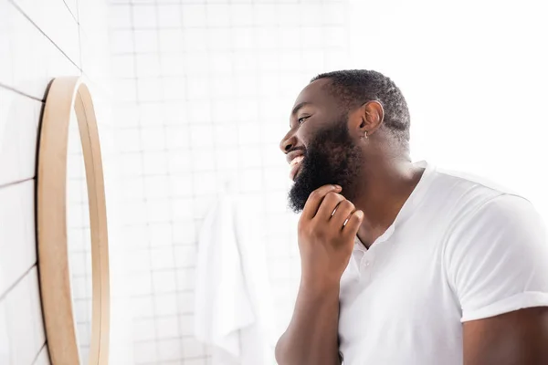 Vue latérale de l'homme afro-américain heureux regardant dans le miroir et touchant barbe — Photo de stock