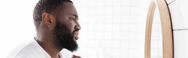 Plano panorámico de hombre afro-americano infeliz mirando la barba en el espejo - foto de stock