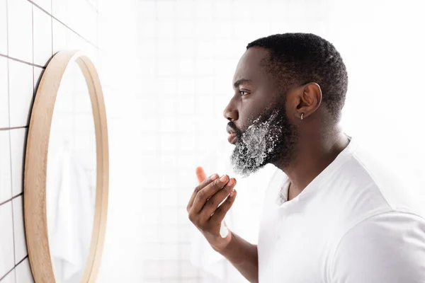 Vista lateral del hombre afroamericano aplicando espuma de afeitar y mirándose en el espejo — Stock Photo