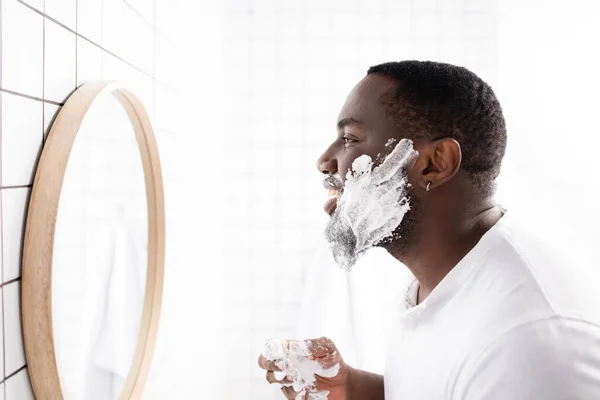 Vista lateral del hombre afroamericano aplicando espuma de afeitar y mirándose en el espejo - foto de stock