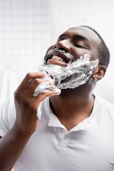 Hombre afro-americano feliz con los ojos cerrados aplicando espuma de afeitar - foto de stock