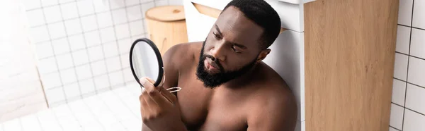 Panoramic shot of afro-american man sitting on bathroom floor and looking at face in small mirror — Stock Photo