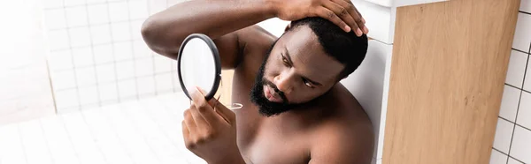 Plano panorámico del hombre afroamericano sentado en el piso del baño y la fijación de pelo en un pequeño espejo - foto de stock