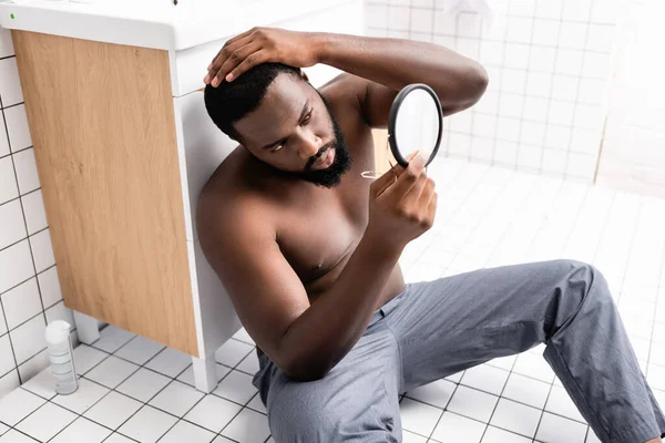 Афро-американец, сидящий на полу в ванной комнате и фиксирующий волосы в маленьком зеркале — стоковое фото