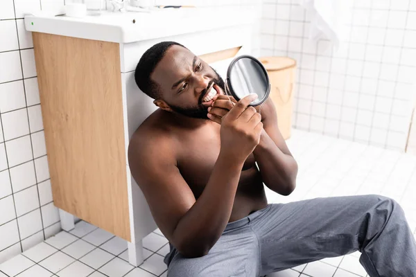 Hombre afroamericano sentado en el piso del baño y mirando chicle en un pequeño espejo — Stock Photo