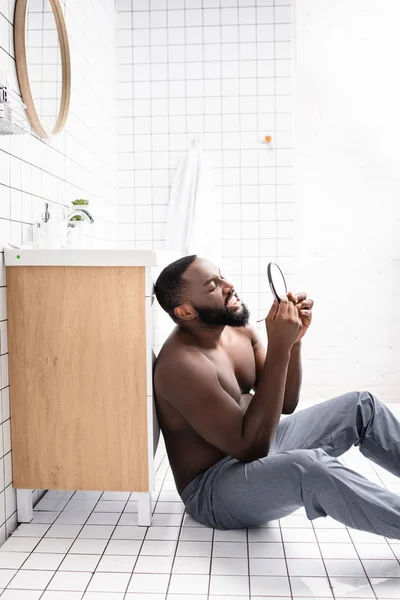 Vista laterale di uomo afro-americano seduto sul pavimento del bagno e guardando gomma in piccolo specchio — Foto stock