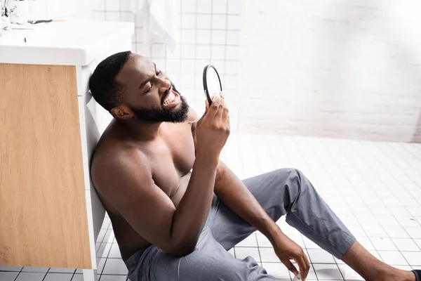 Afro-american man sitting on bathroom floor and looking at gum in small mirror — Stock Photo