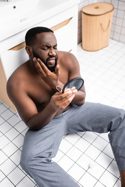 Hombre afroamericano sentado en el suelo del baño y haciendo muecas de dolor de muelas — Stock Photo