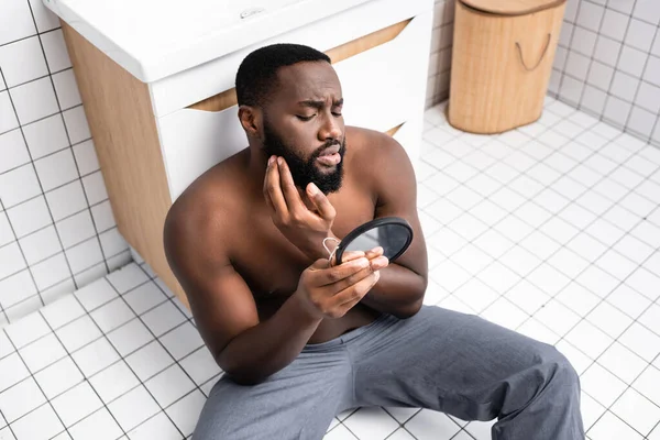 Afro-américain assis sur le sol de la salle de bain et fixant les cheveux sur la barbe — Photo de stock