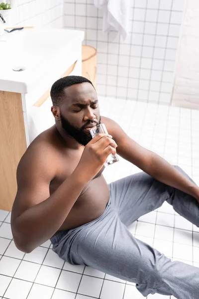 Hombre afroamericano sentado en el suelo del baño y agua potable — Stock Photo