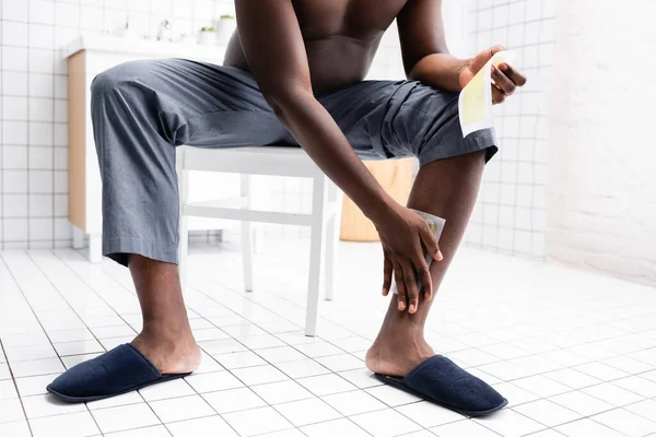 Vista recortada del hombre afroamericano aplicando tiras de cera sentado en el baño - foto de stock