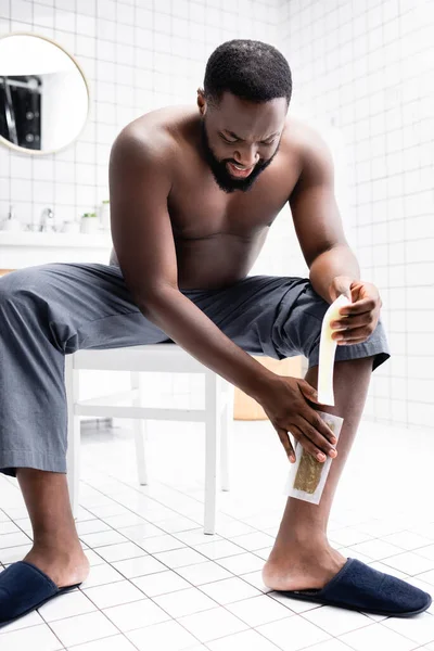 Hombre afroamericano aplicando tiras de cera sentado en el baño - foto de stock