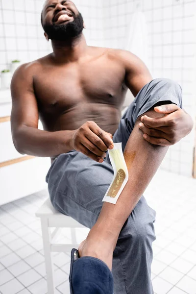 Hombre afroamericano arrancando tiras de cera y sufriendo de dolor - foto de stock