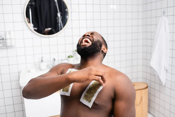 Hombre afroamericano arrancando tiras de cera y gritando de dolor - foto de stock