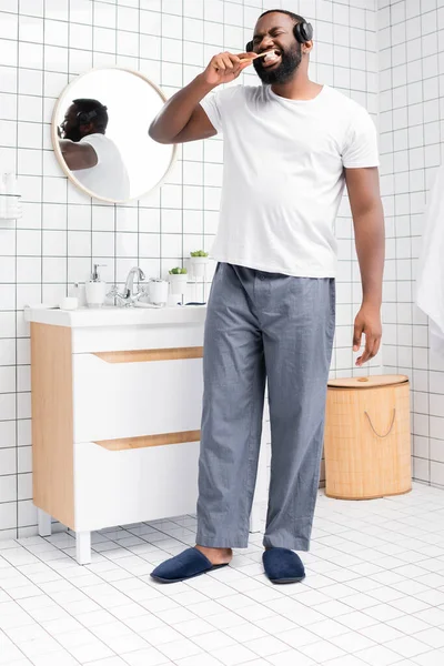Longitud completa del hombre afroamericano con auriculares cepillándose los dientes - foto de stock
