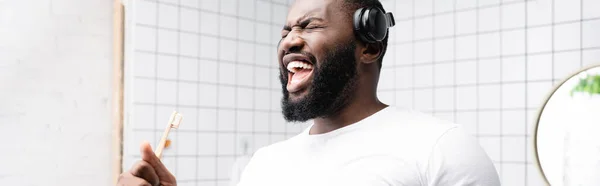 Tiro panorâmico de homem afro-americano com fones de ouvido cantando em escova de dentes — Fotografia de Stock