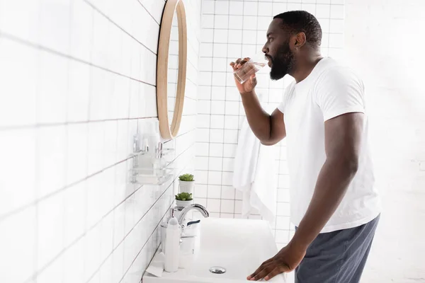 Vista lateral do homem afro-americano água potável e olhando no espelho — Fotografia de Stock