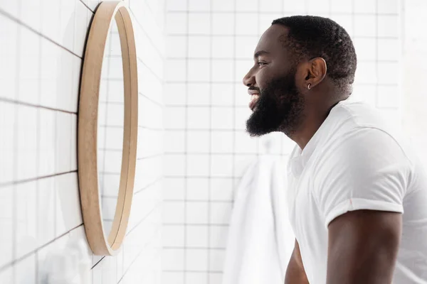 Vista lateral del hombre afroamericano sonriendo y mirándose en el espejo - foto de stock