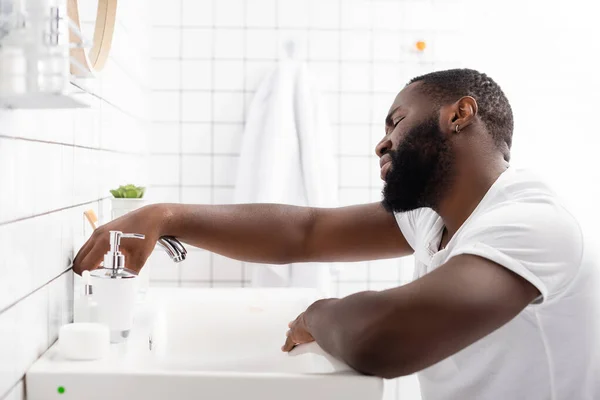 Cansado afro-americano homem inclinado no dissipador e tentando aberto torneira — Fotografia de Stock